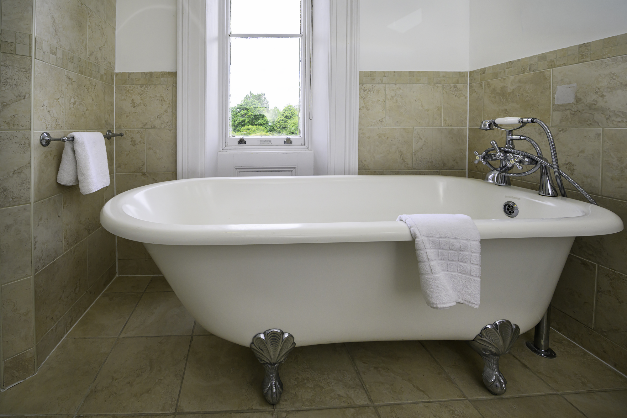A vintage clawfoot bathtub in a bathroom with beige tile walls and flooring, a towel hanging on the side, and a window revealing greenery outside.