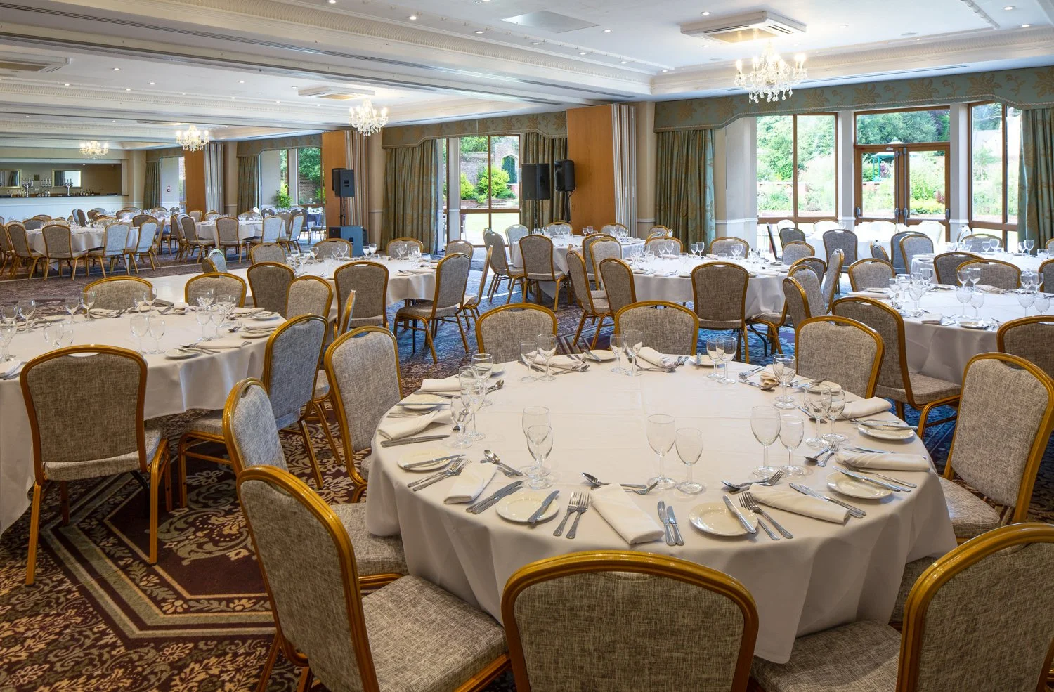 Elegant banquet hall set up with round tables covered in white tablecloths, decorated with glassware, silverware, and napkins, with large windows and chandeliers overhead.