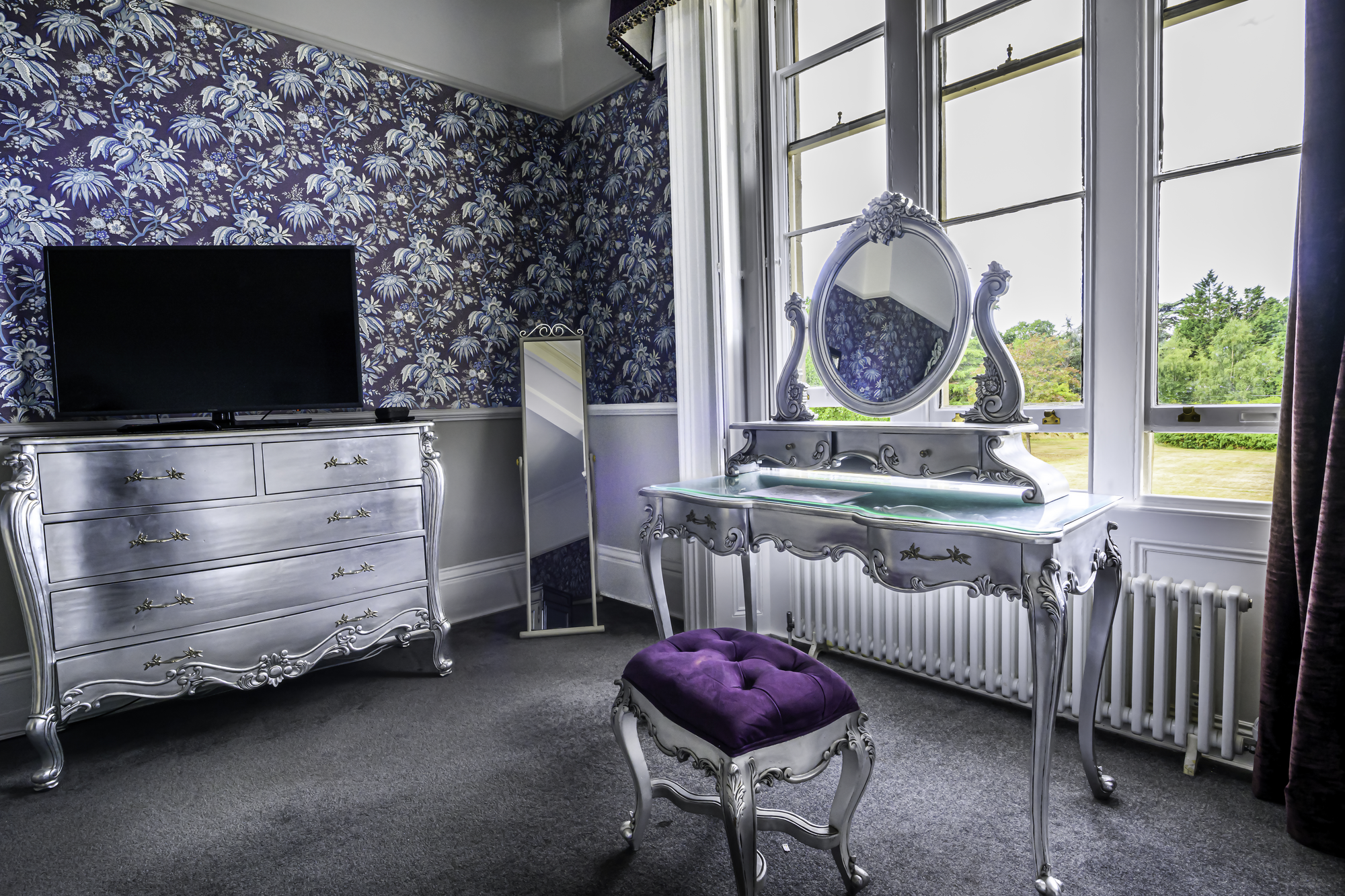 A bedroom featuring a silver ornate vanity with a mirror, a matching silver dresser with a TV on top, a purple velvet stool, a full-length mirror, and a window with scenic greenery outside.