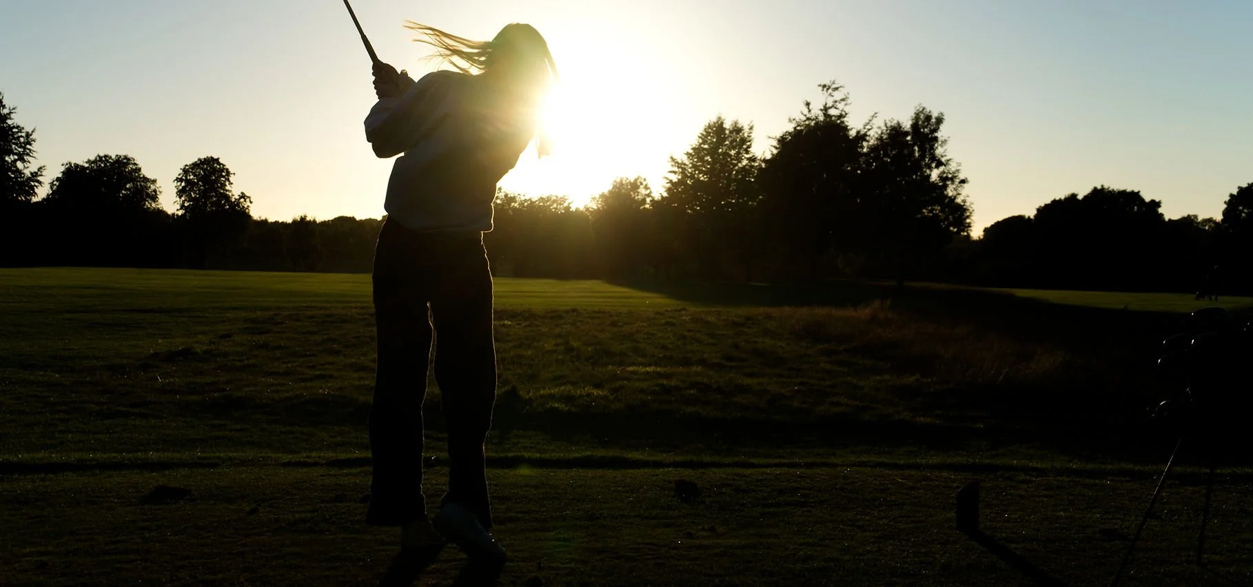 A person playing golf, swinging a club on a golf course during sunset with trees in the background.