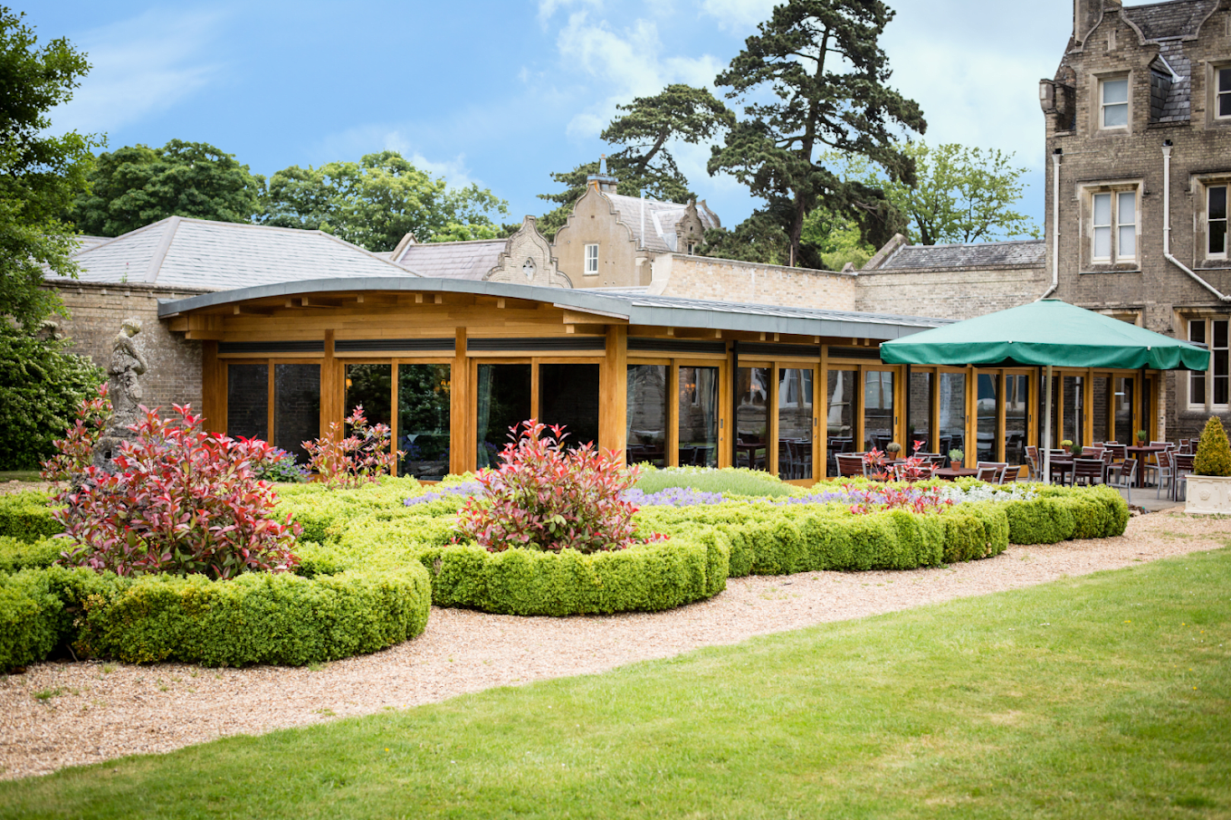 An outdoor patio with wooden and glass walls, a green umbrella covering some tables and chairs, surrounded by landscaped garden with bushes, flowers, and trees, in front of a stone building.