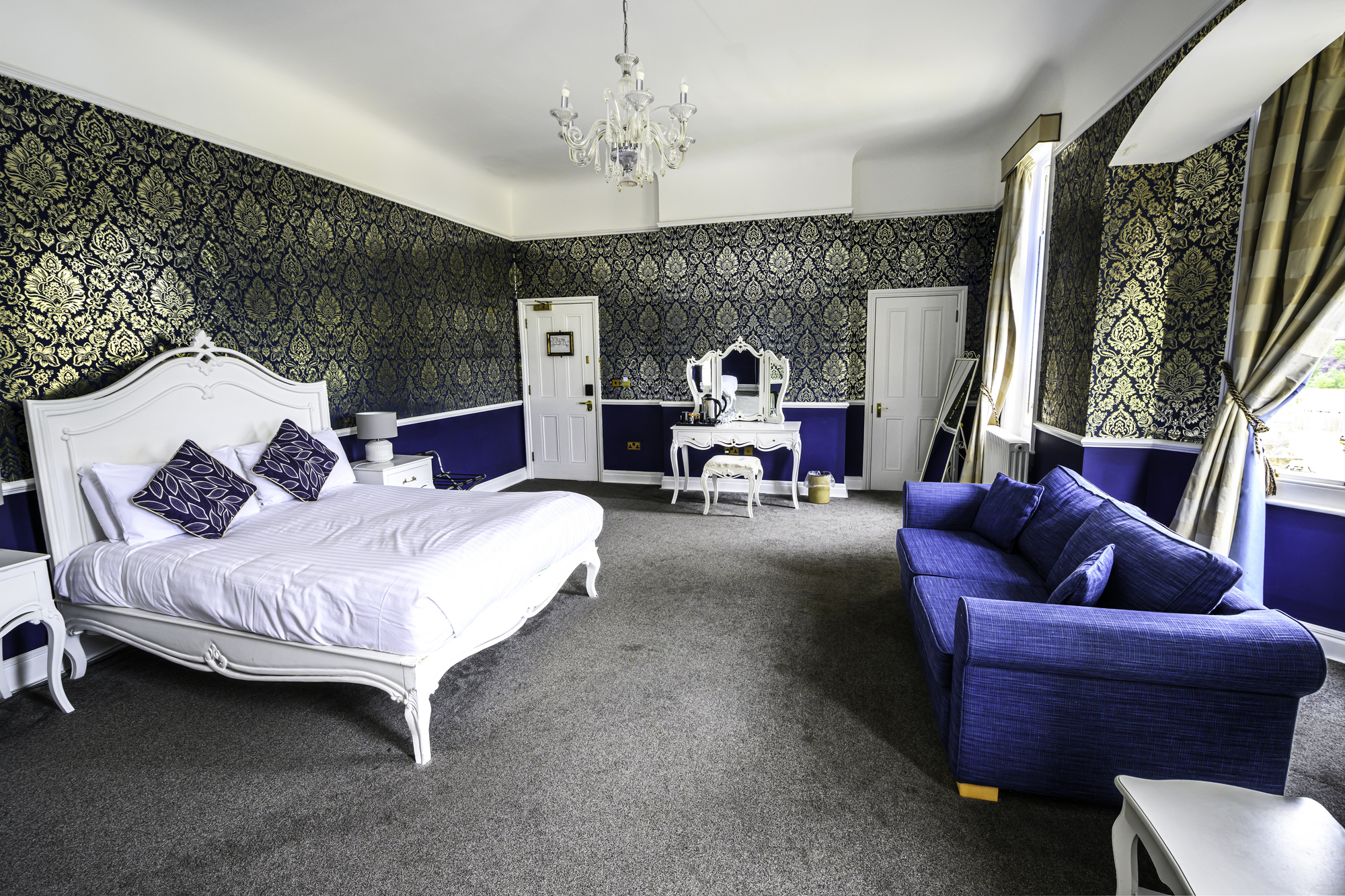 Elegant bedroom with white bed, blue velvet sofa, patterned wallpaper, chandelier, and large windows with curtains.