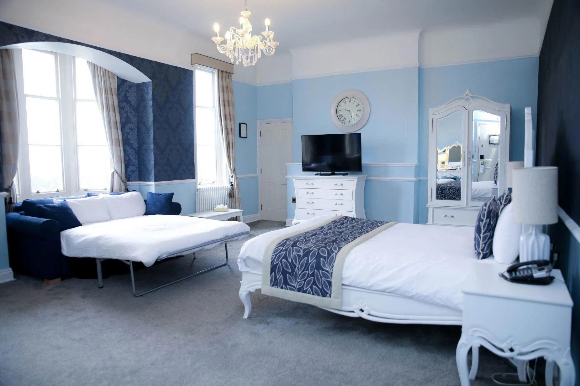 A spacious, well-lit hotel room with a bed, a blue sofa, a dresser with a TV, and decorative blue and white accents.