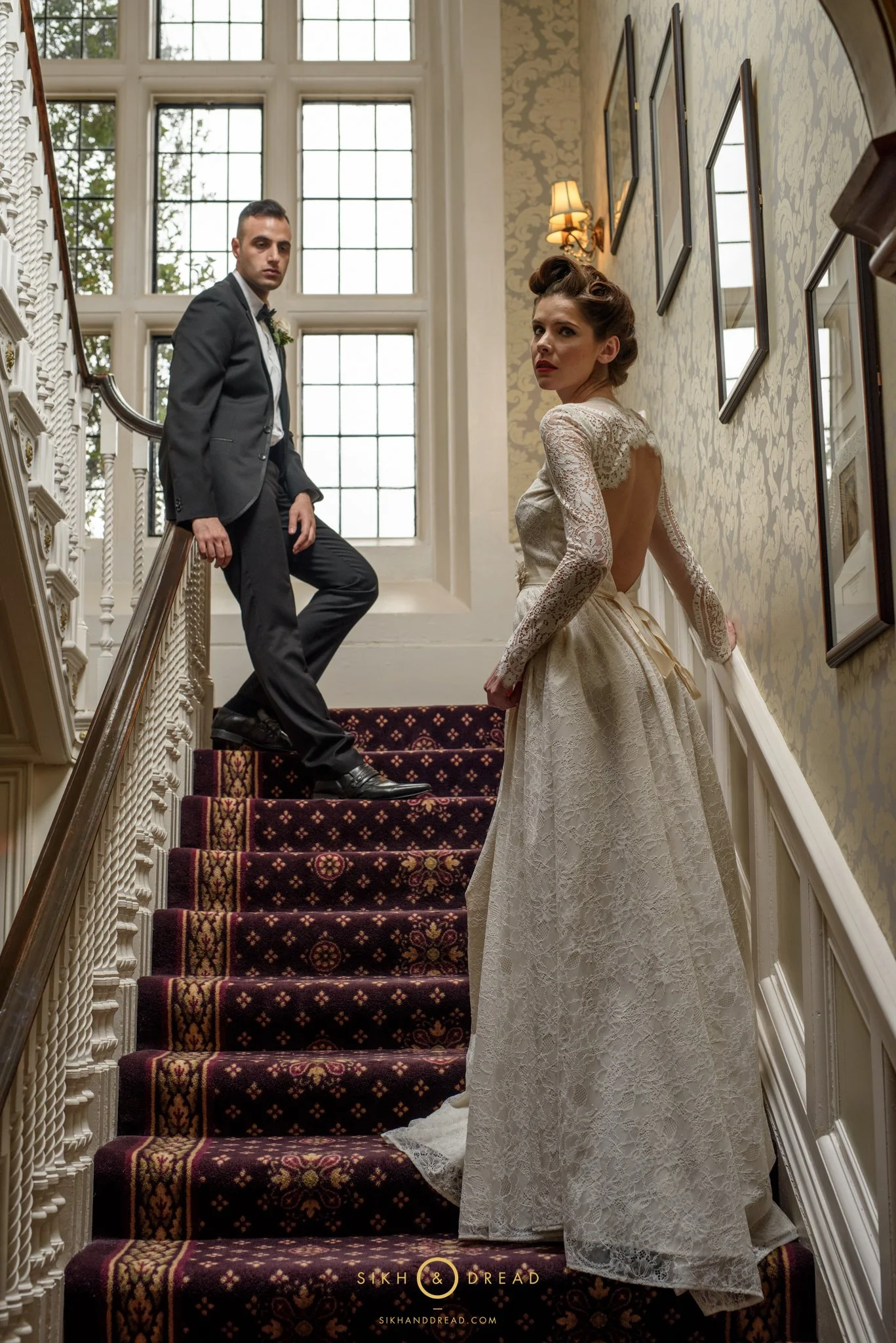 A woman in a vintage lace wedding dress standing on a staircase, looking back, with a man in a tuxedo nearby at the top of the staircase.