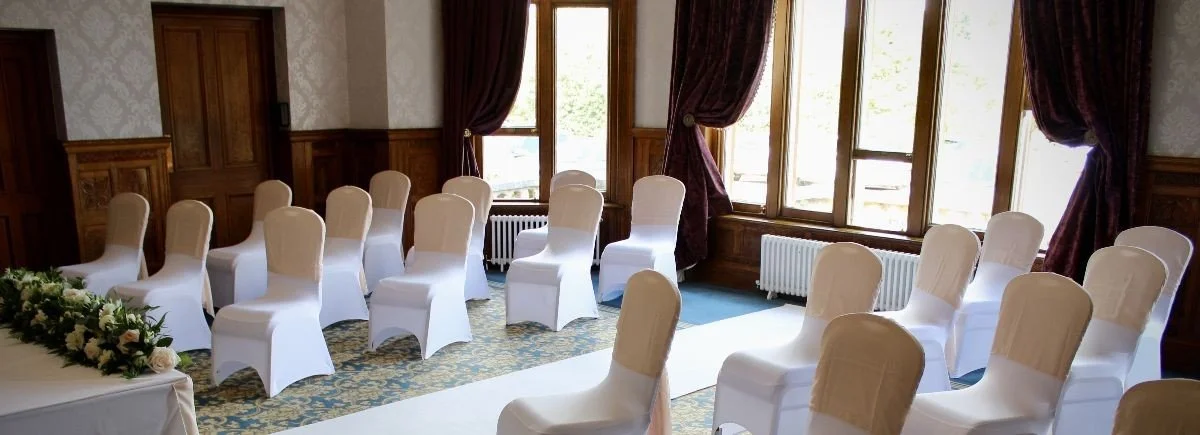 Empty chairs arranged for a wedding ceremony in a decorated room with large windows and purple curtains.