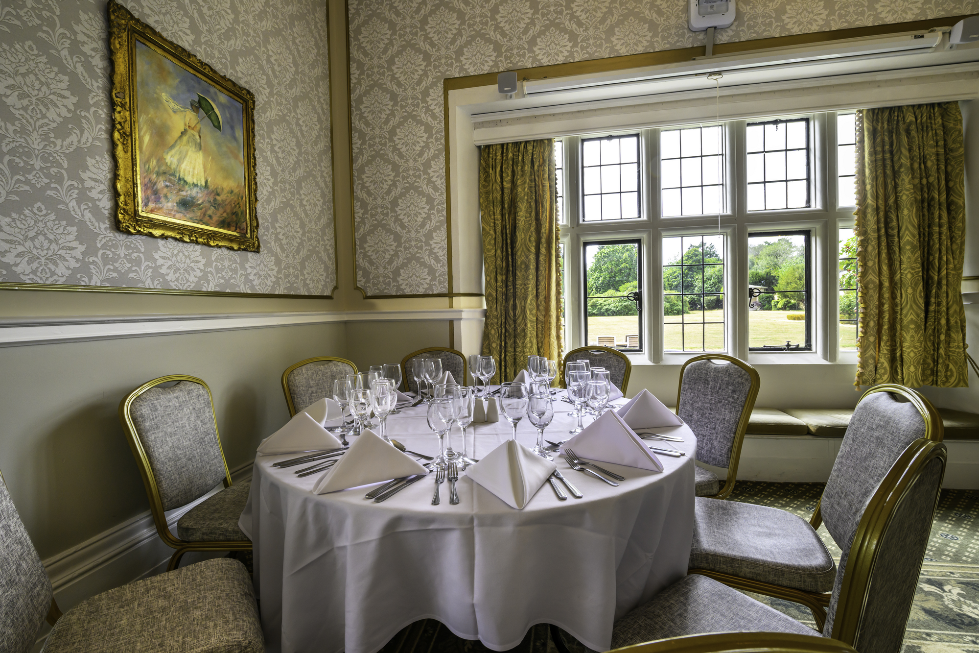 A round dining table set with white linen, folded napkins, and glassware in a bright room with large windows, patterned wallpaper, and framed painting.