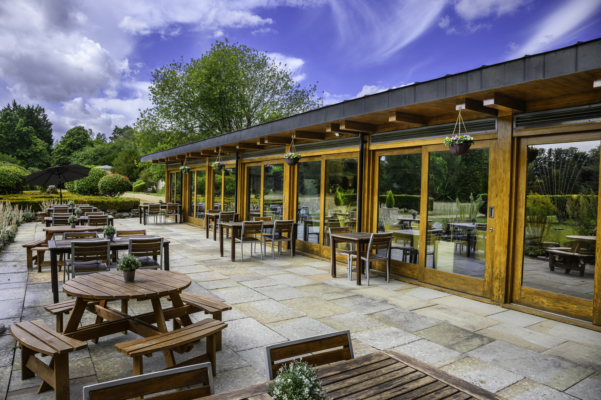 Outdoor patio with wooden tables and chairs, some with small potted plants, set in front of a building with large glass windows and hanging flower pots, surrounded by lush greenery and trees under a partly cloudy sky.