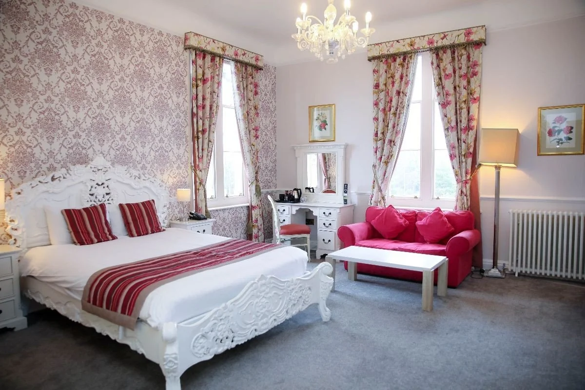 A bedroom with a white ornate bed, red and white striped pillows, floral curtains, a pink sofa, a white table, a writing desk with a mirror, and a chandelier.