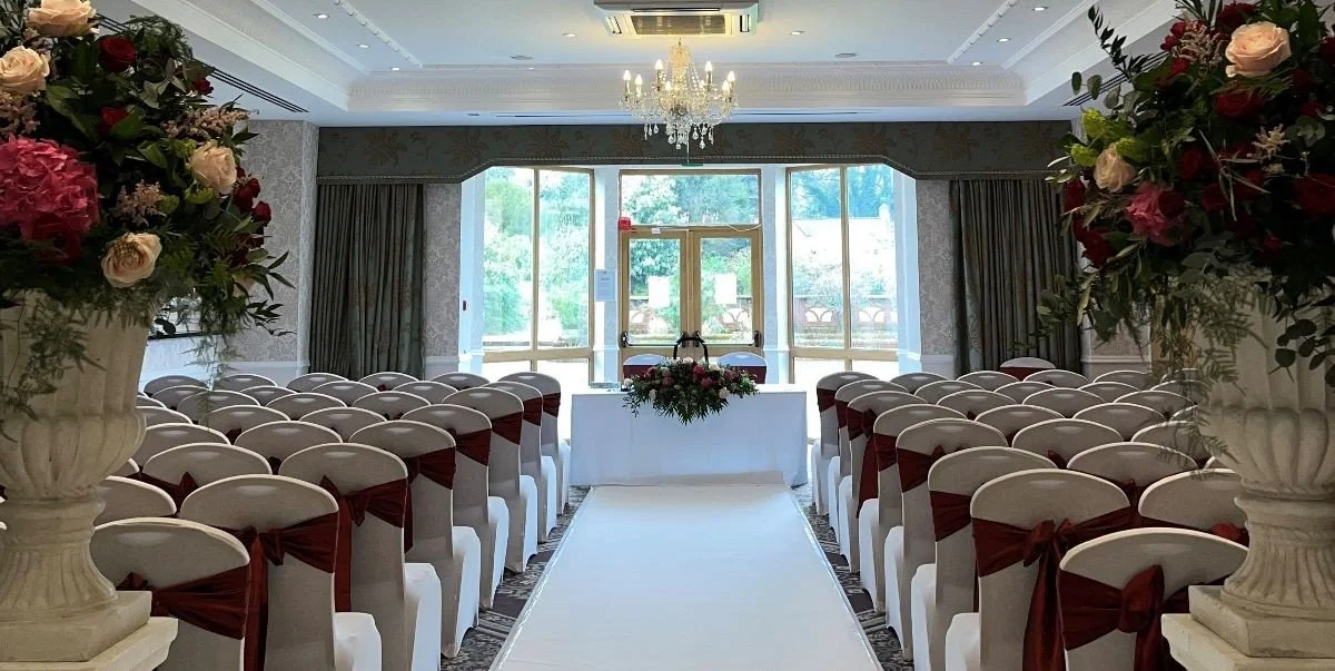 Indoor wedding ceremony setup with rows of white chairs with burgundy sashes facing an altar with floral arrangements, large vases of flowers on pedestals, and a chandelier, in front of glass doors and windows with outdoor greenery visible.