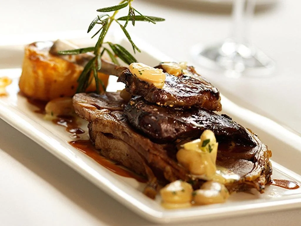A plated serving of cooked lamb chops garnished with herbs, garlic, and butter, served with roasted potatoes.