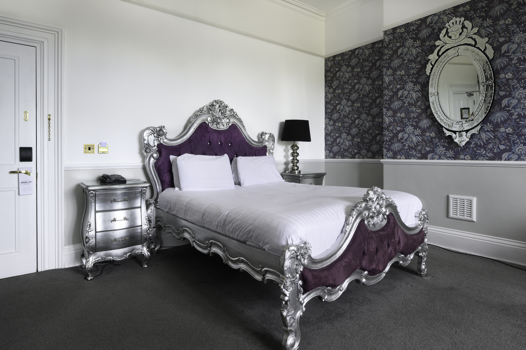 A luxurious hotel bedroom with a large ornate silver and purple bed, two matching silver nightstands, a black lamp on the right side, and a decorative mirror on a dark floral wallpapered wall.