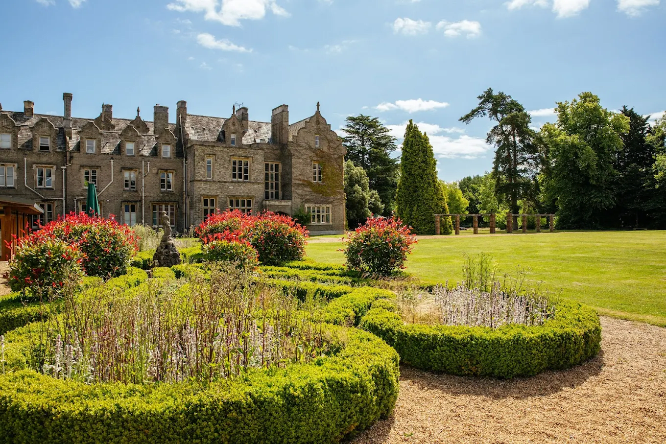 A historic stone mansion with a well-kept garden featuring colorful flowers and lush green trees under a partly cloudy sky.