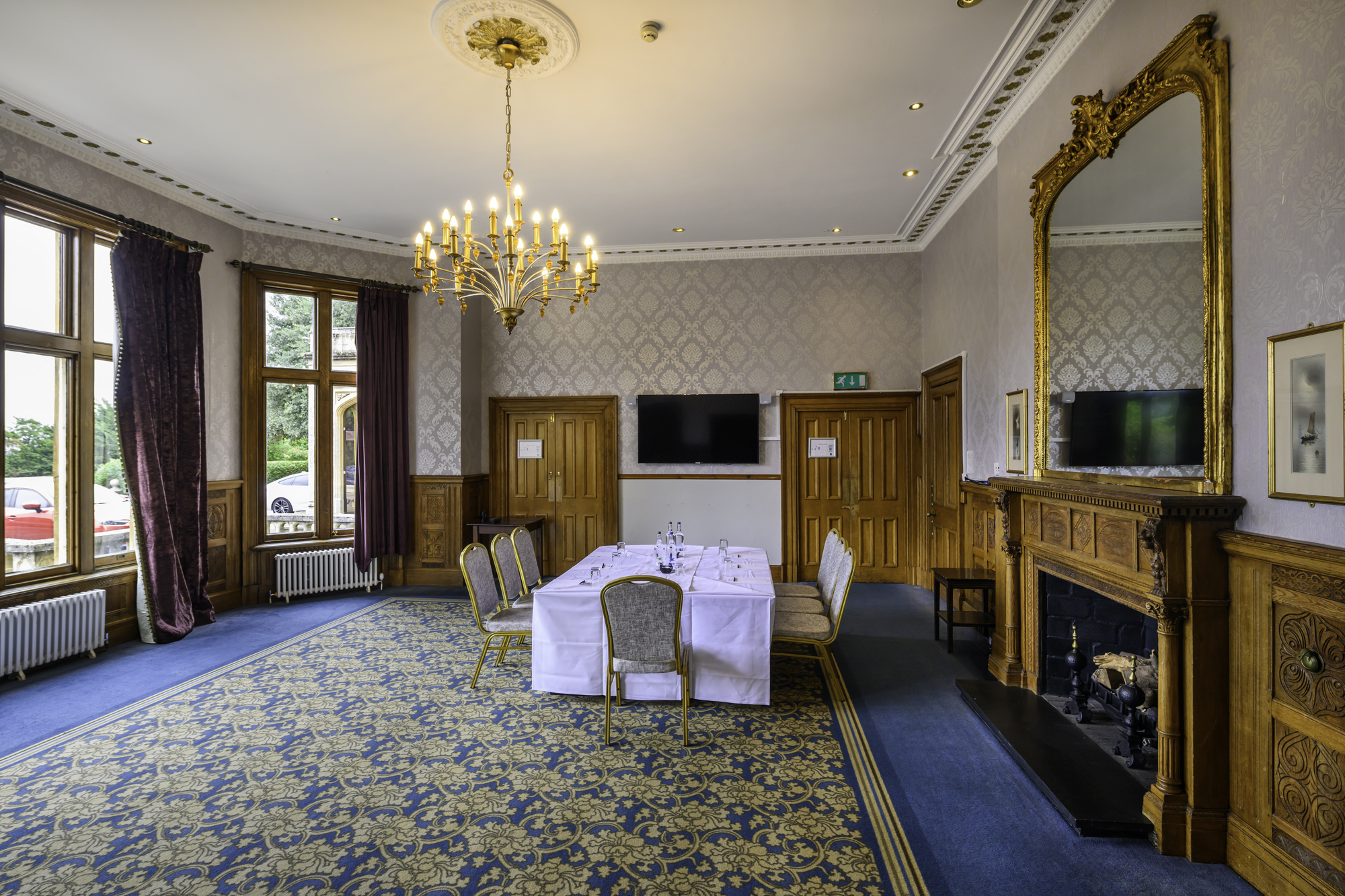Elegant dining room with large windows, blue patterned carpet, chandelier, fireplace, and a table set for a formal meal.