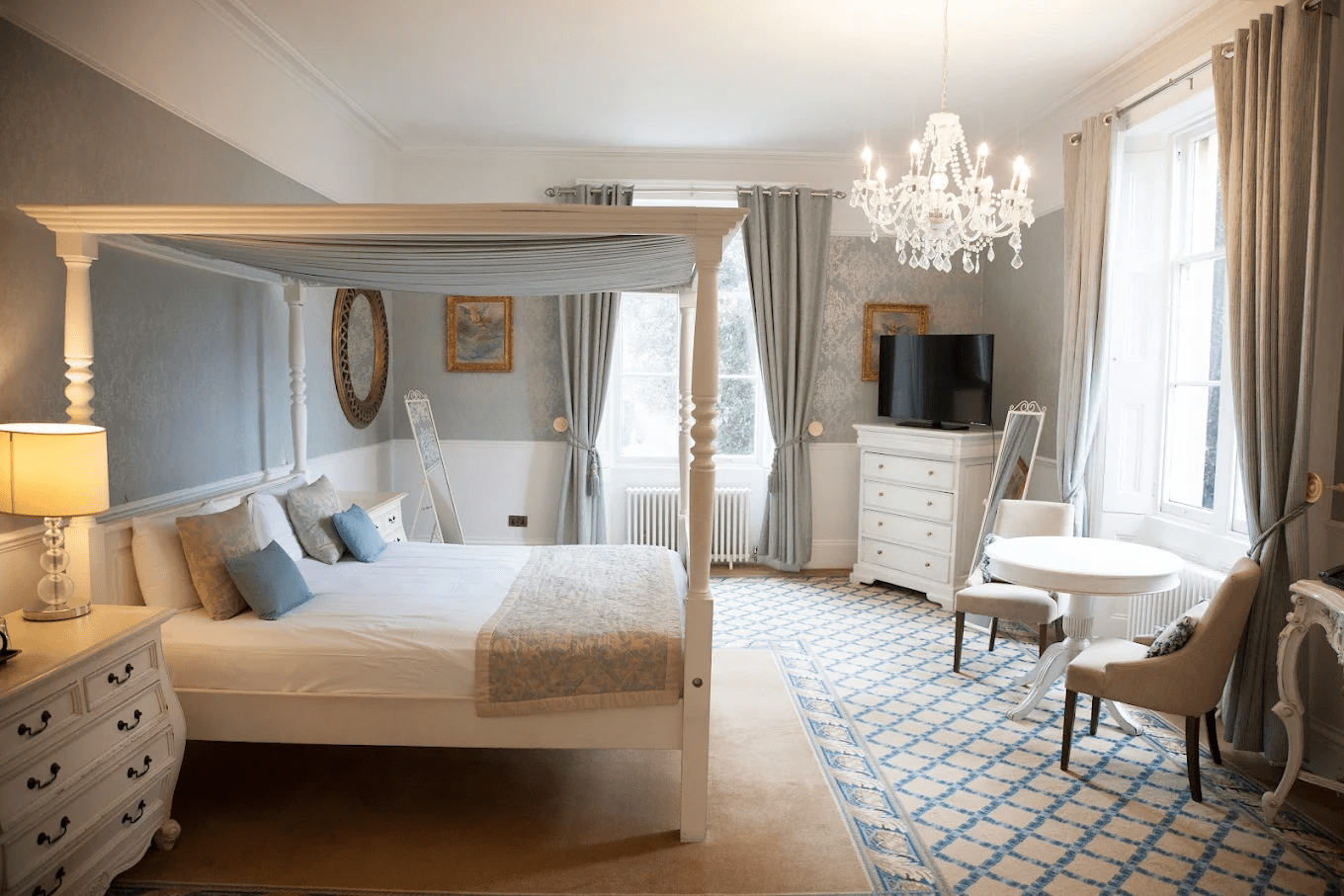 Elegant bedroom with a four-poster bed, chandelier, dresser with a flat-screen TV, round table, and upholstered chairs, decorated in soft blue and white tones with large windows and curtains.