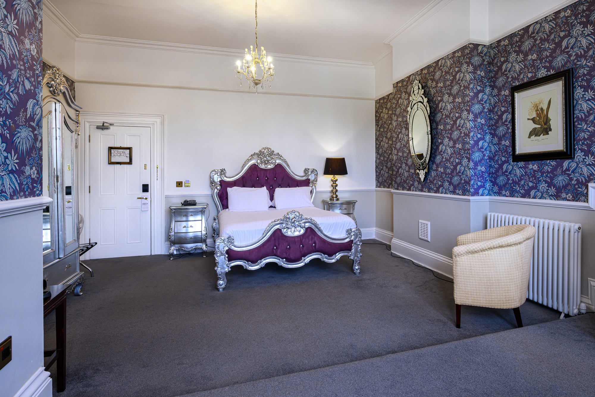 Elegant bedroom with a purple velvet and silver ornate bed, white bedding, silver side tables, a black and gold lamp, a cream upholstered chair, framed artwork, a mirror, a chandelier, and dark floral wallpaper on one wall.