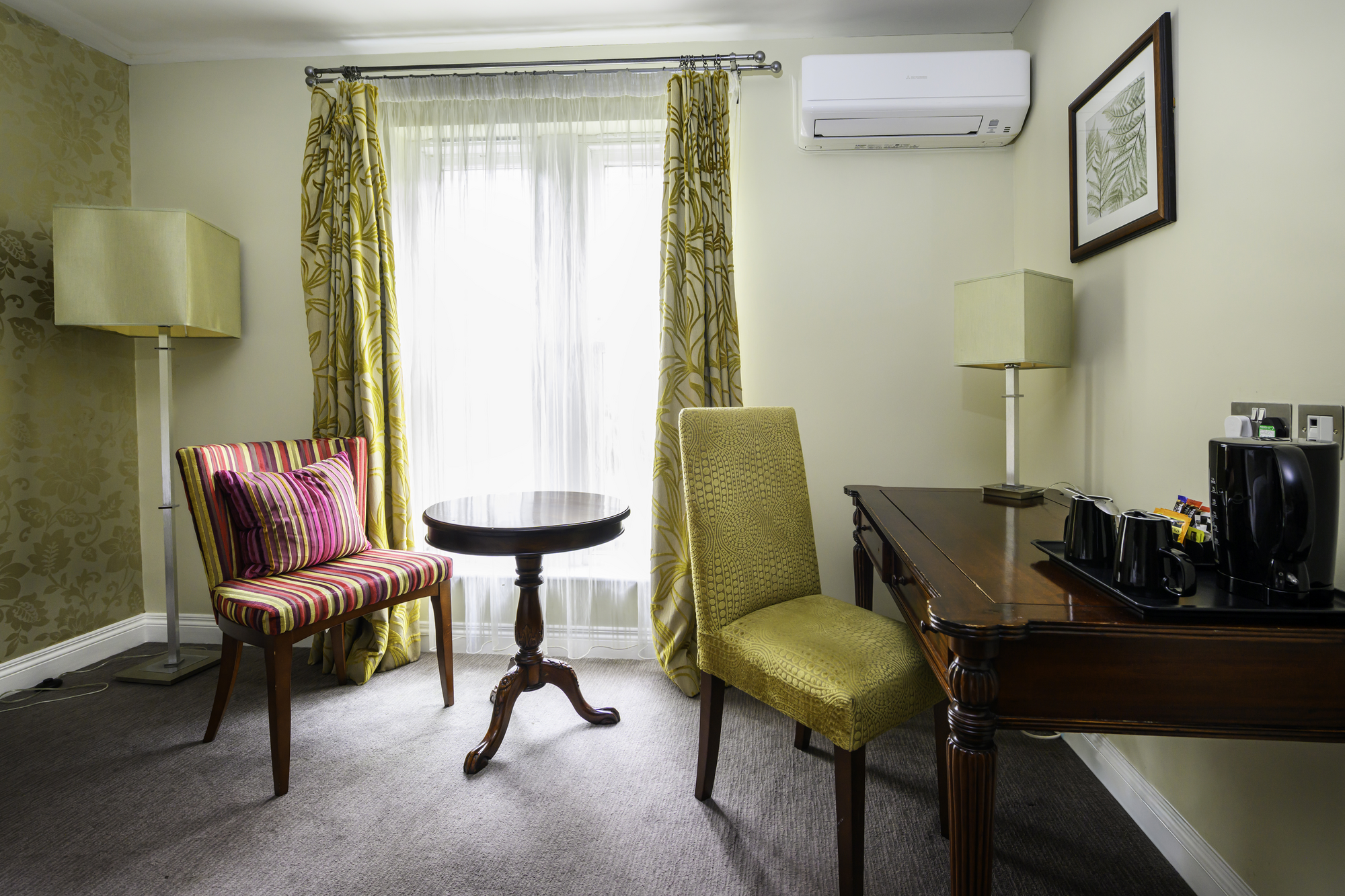 Hotel room with yellow accents, a small round table, a striped armchair with a pillow, a green chair, a wooden desk with a tea and coffee setup, framed botanical art on the wall, yellow patterned curtains, and an air conditioning unit.