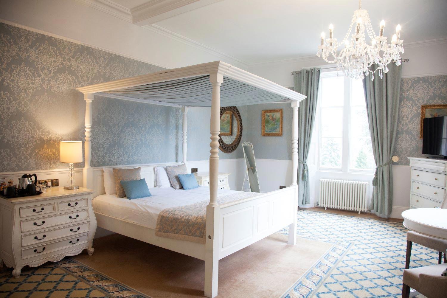 Elegant bedroom with a white four-poster bed, blue and white decor, a chandelier, a window with curtains, a dresser with a television, and artwork on the walls.