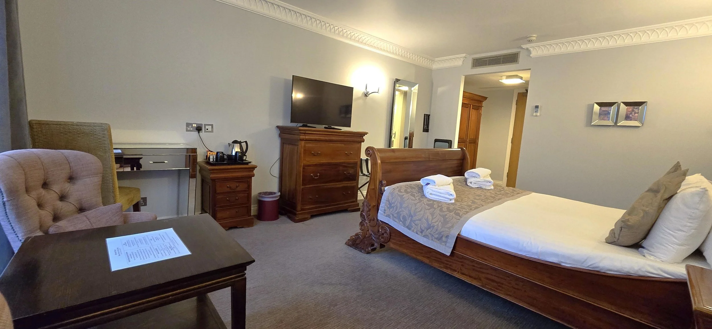 A hotel room with a large wooden bed, white linens, and folded towels on top. There is a beige armchair, a wooden table, and a dresser with a flat-screen TV. The room has light-colored walls, crown molding, and framed pictures on the wall.