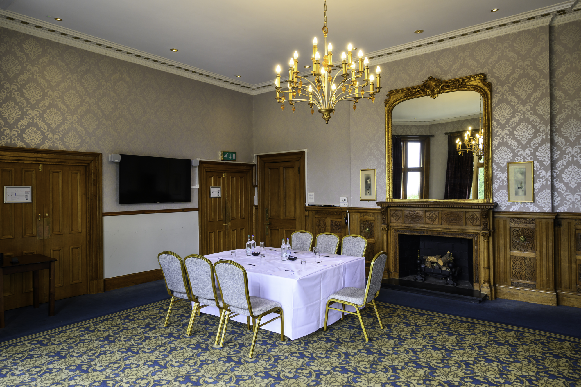 A formal dining room with a chandelier, a large mirror above a fireplace, a table set with water bottles and glasses, and eight chairs with upholstered seats.