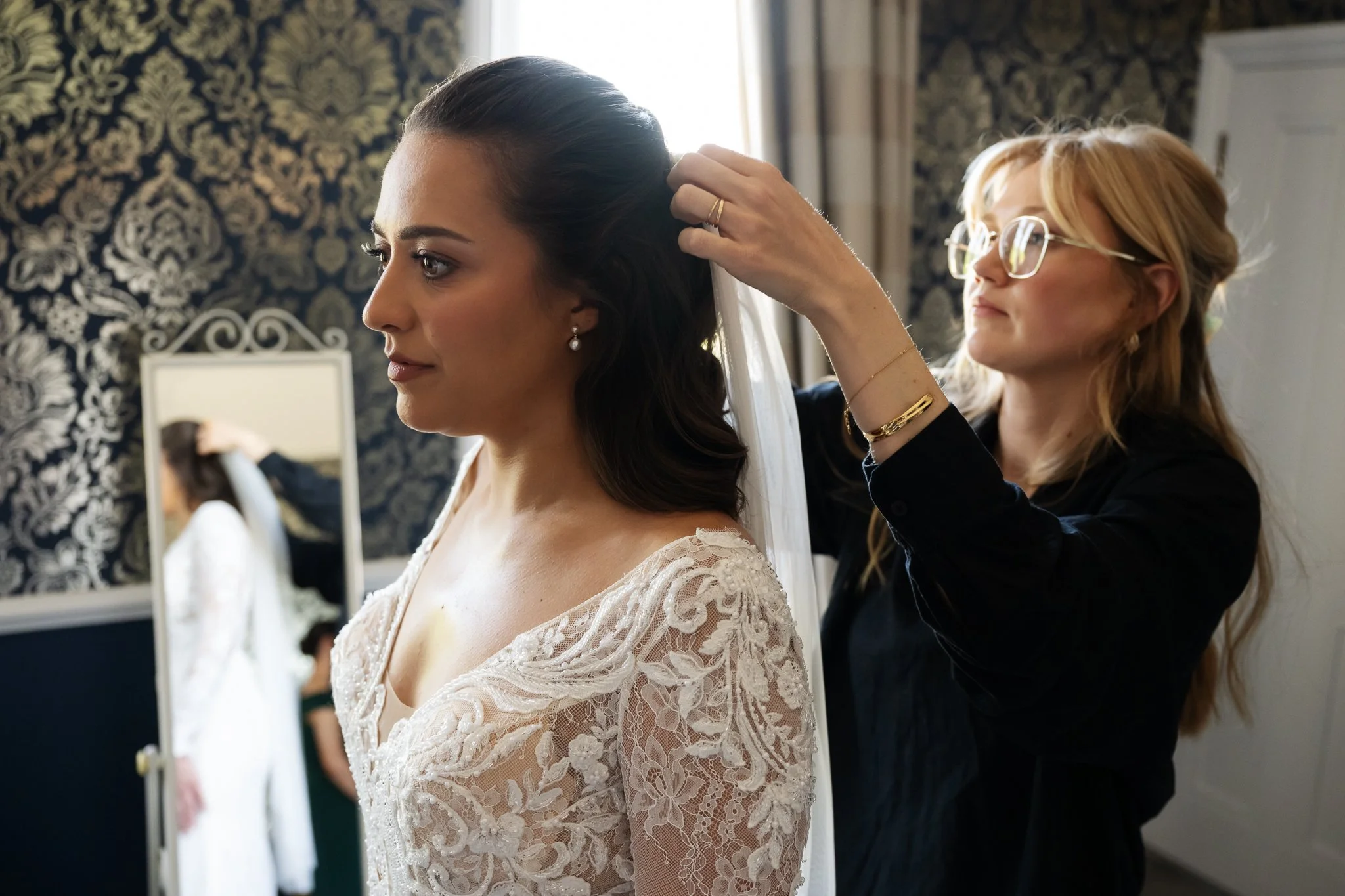 A woman helping a bride with her veil in a room with floral wallpaper and a mirror.