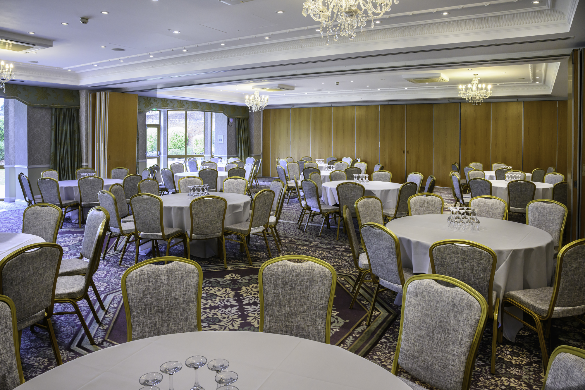 A spacious banquet hall with round tables covered in white tablecloths, set with wine glasses, and surrounded by cushioned chairs with gold frames. Elegant chandeliers hang from the ceiling, and large windows let in natural light. The walls are decorated with patterned wallpaper and wooden panels, and the floor is carpeted with an intricate floral design.