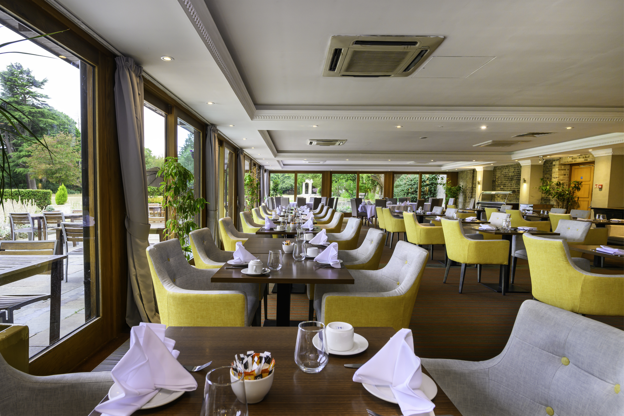 Elegant restaurant dining area with yellow and gray chairs, set tables with white napkins, glassware, and coffee cups, large windows with curtains, green plants, and outdoor patio with tables and chairs visible outside.