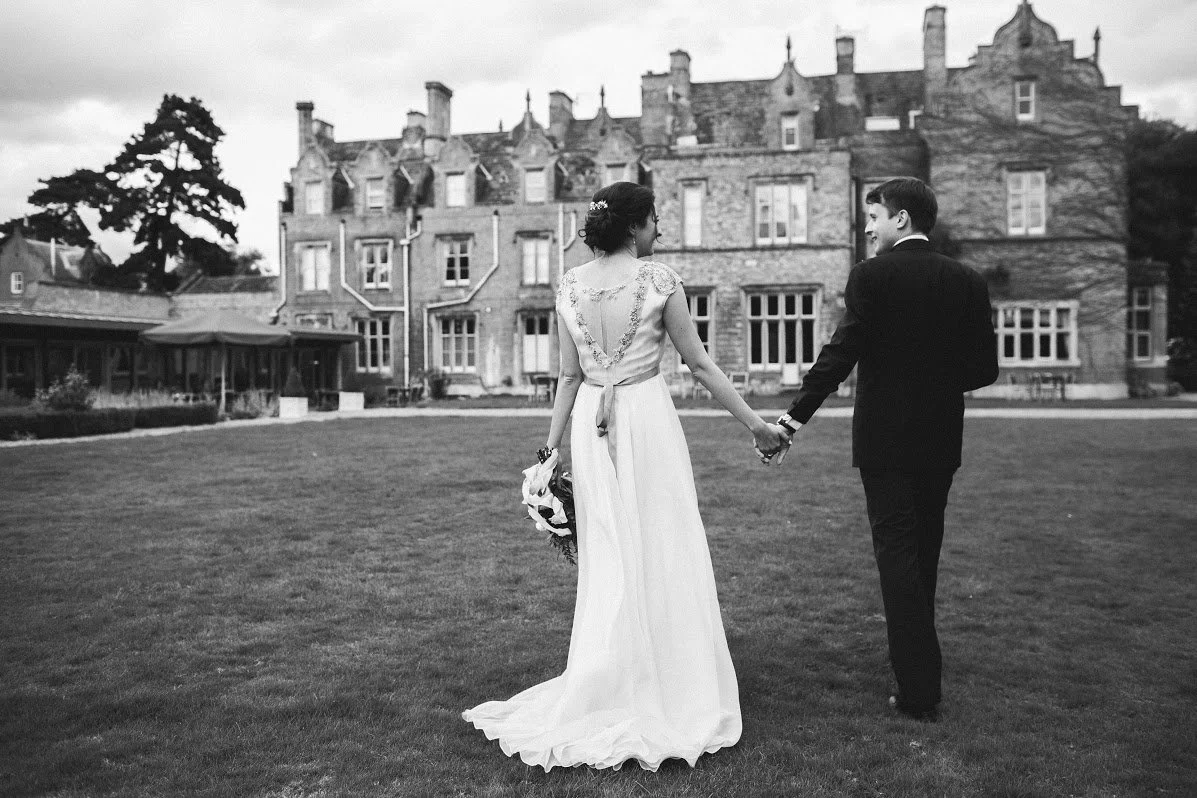 A black-and-white photo of a bride and groom holding hands on a lawn in front of a large historic building, with the bride in a long wedding dress and the groom in a dark suit.