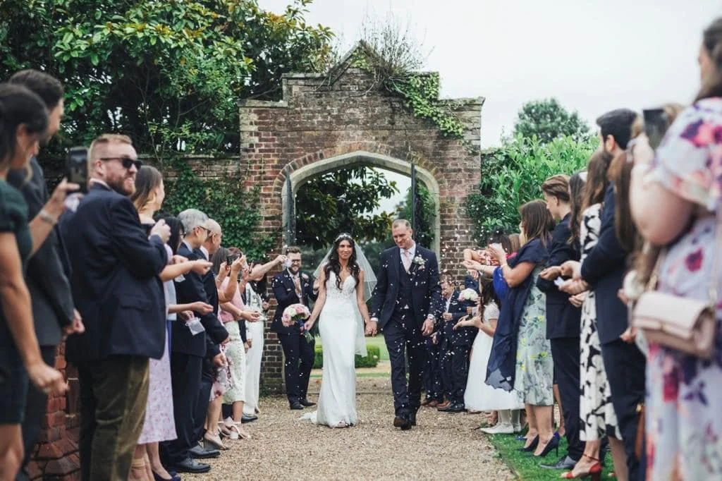 Couple walking hand in hand through a wedding arch surrounded by guests throwing confetti.