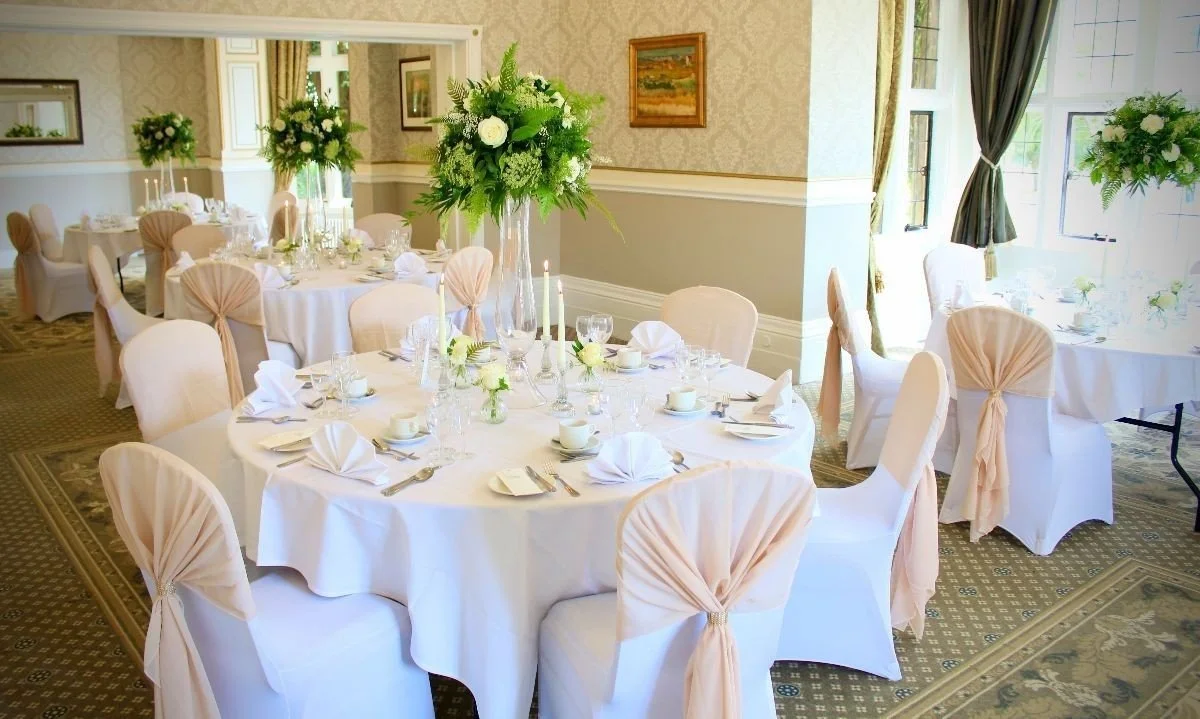 Elegant banquet room decorated for a formal event with round tables covered in white tablecloths, pink chair covers with bows, and tall floral centerpieces with white roses and greenery. The room has large windows with curtains, framed artwork on the