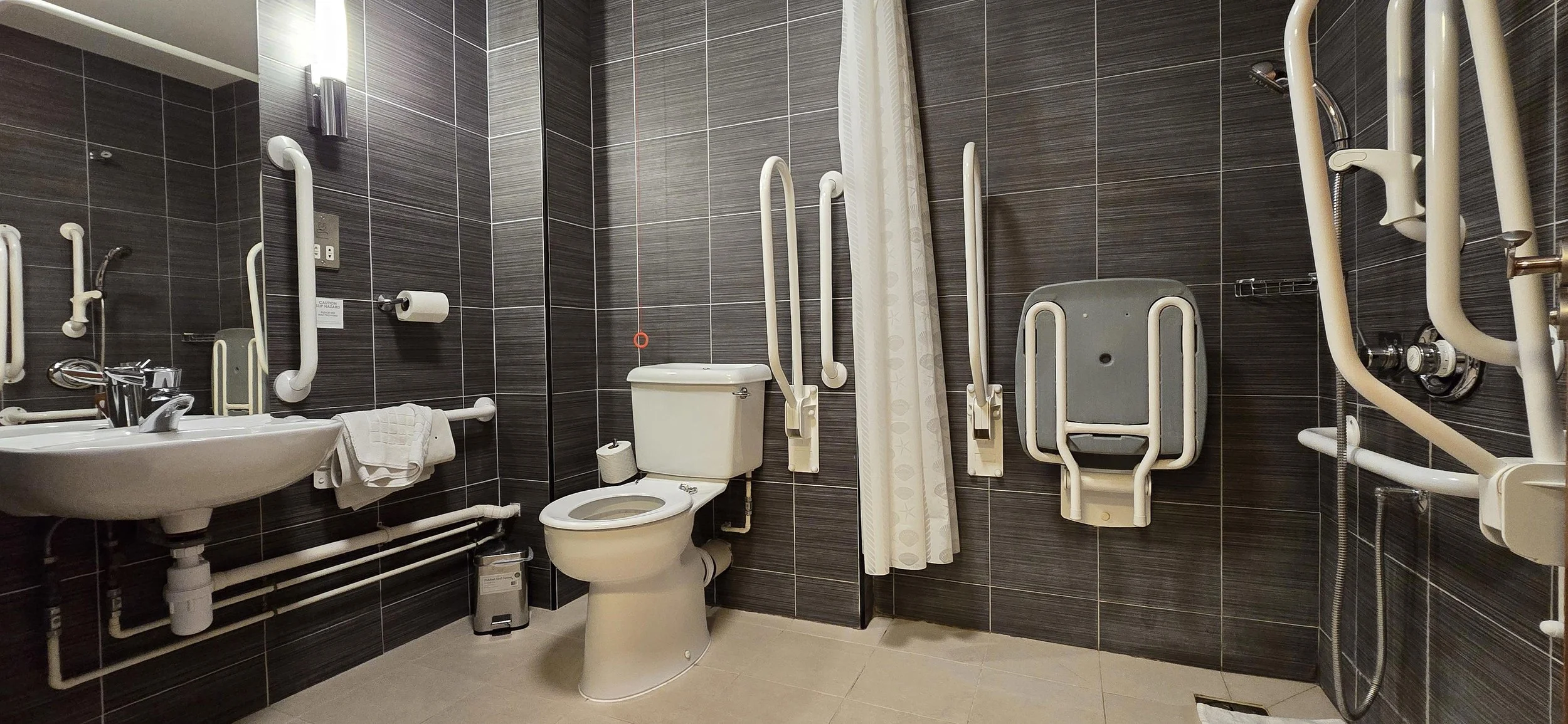 A bathroom with dark tiled walls and beige tiles on the floor, equipped with accessibility features including grab bars around the toilet, a fold-down shower seat, and grab bars around the sink and shower area, with a curtain partially covering the s