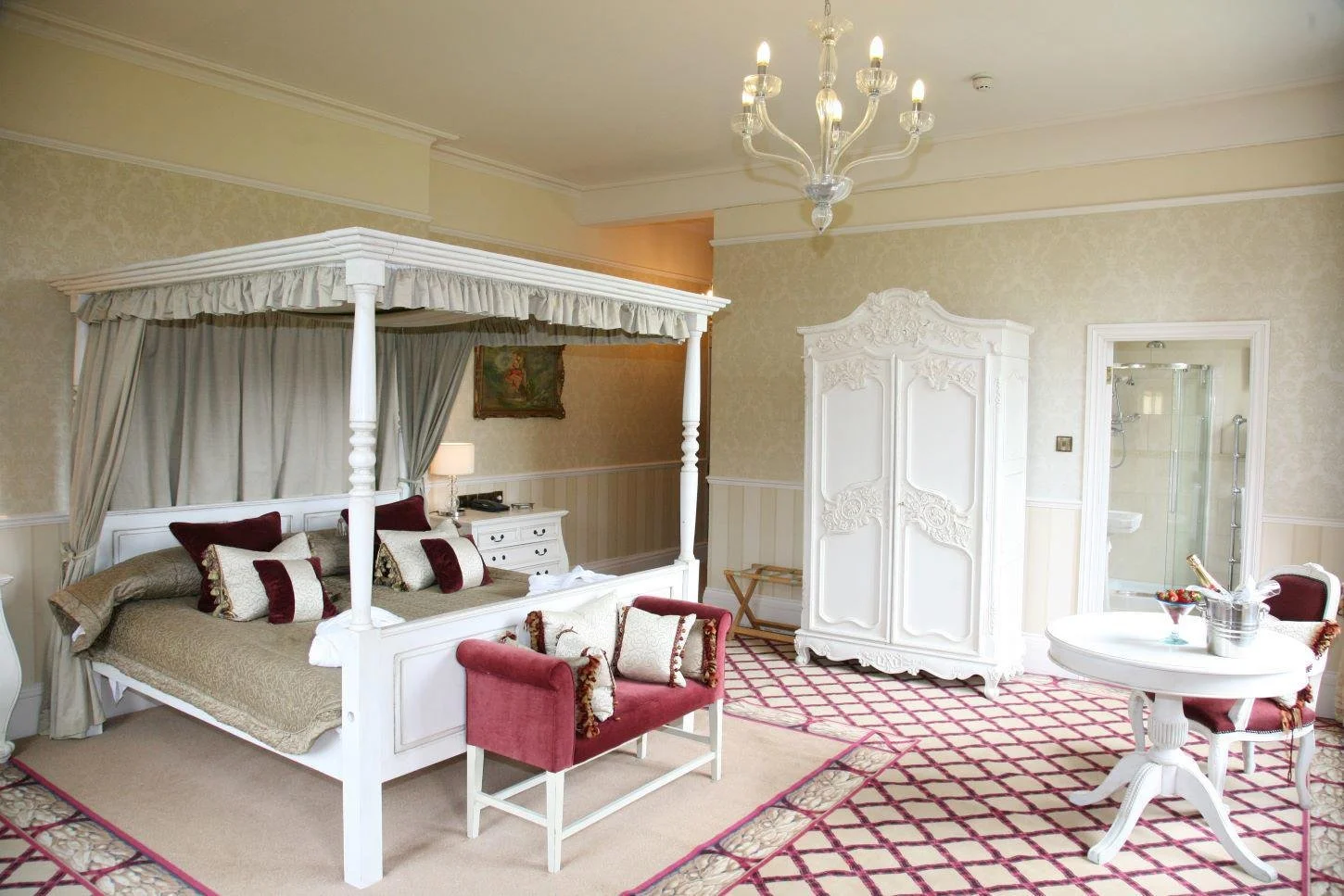 Elegant hotel room with four-poster bed, red and white cushioned furniture, ornate white armoire, chandelier, and a glass-enclosed bathroom.