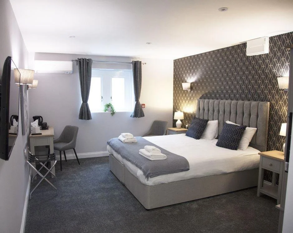 A modern hotel room with a large bed, nightstands, and lamps, a window with curtains, a small desk area, and a wall-mounted TV.