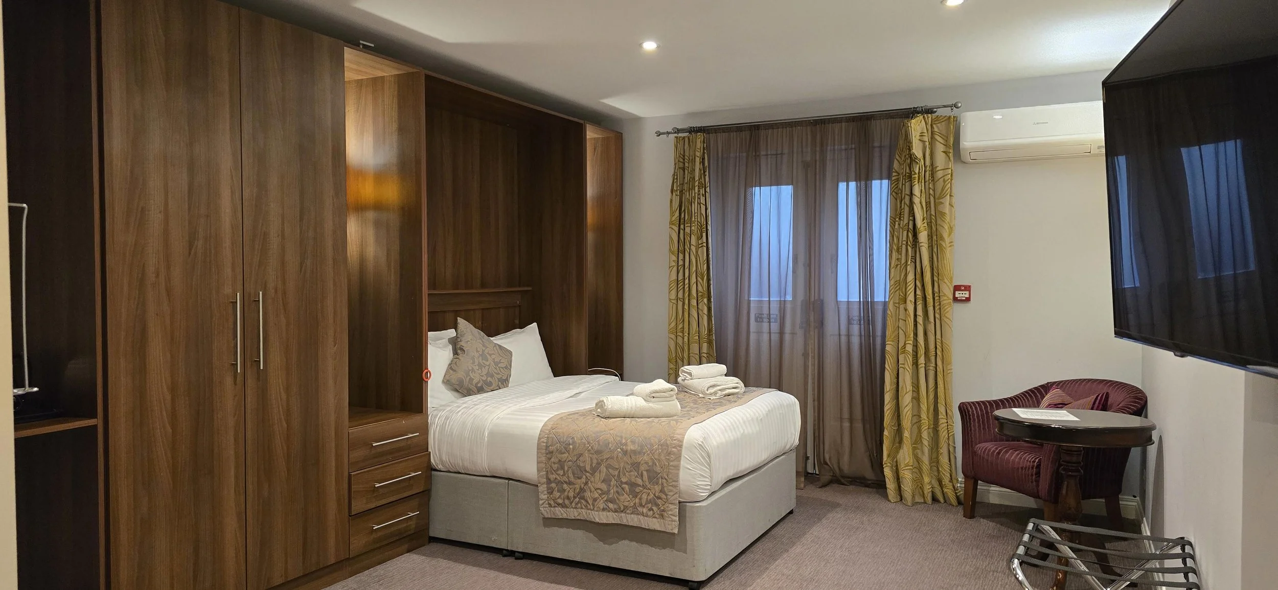 Hotel room with a bed, wooden wardrobe, window with curtains, a small table with chair, and a wall-mounted TV.