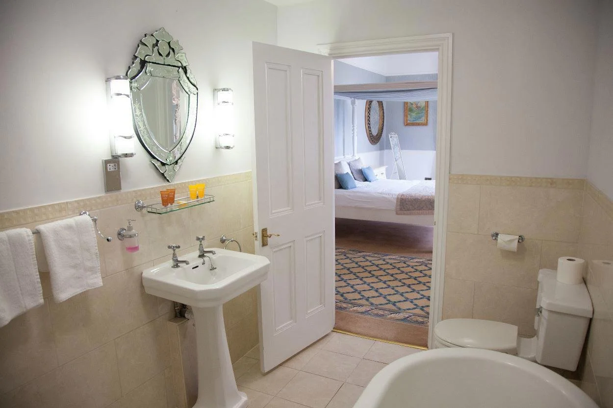 A bathroom with beige tiled walls and floor, a white pedestal sink, a mirror, wall-mounted lights, and a toilet. There are towels hanging on a towel rack, and the bathroom door is open, leading to a bedroom with a bed, blue pillows, and a rug.