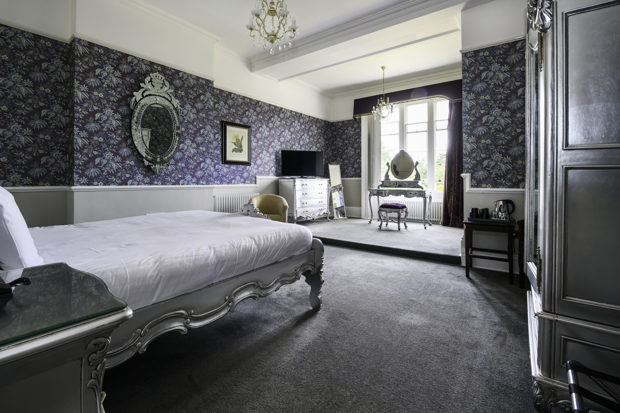 A luxurious vintage bedroom with ornate silver furniture, purple and floral patterned wallpaper, a large white bed, a chandelier, a dresser, a seating area by large windows, a mirror, and a black side table with a kettle.