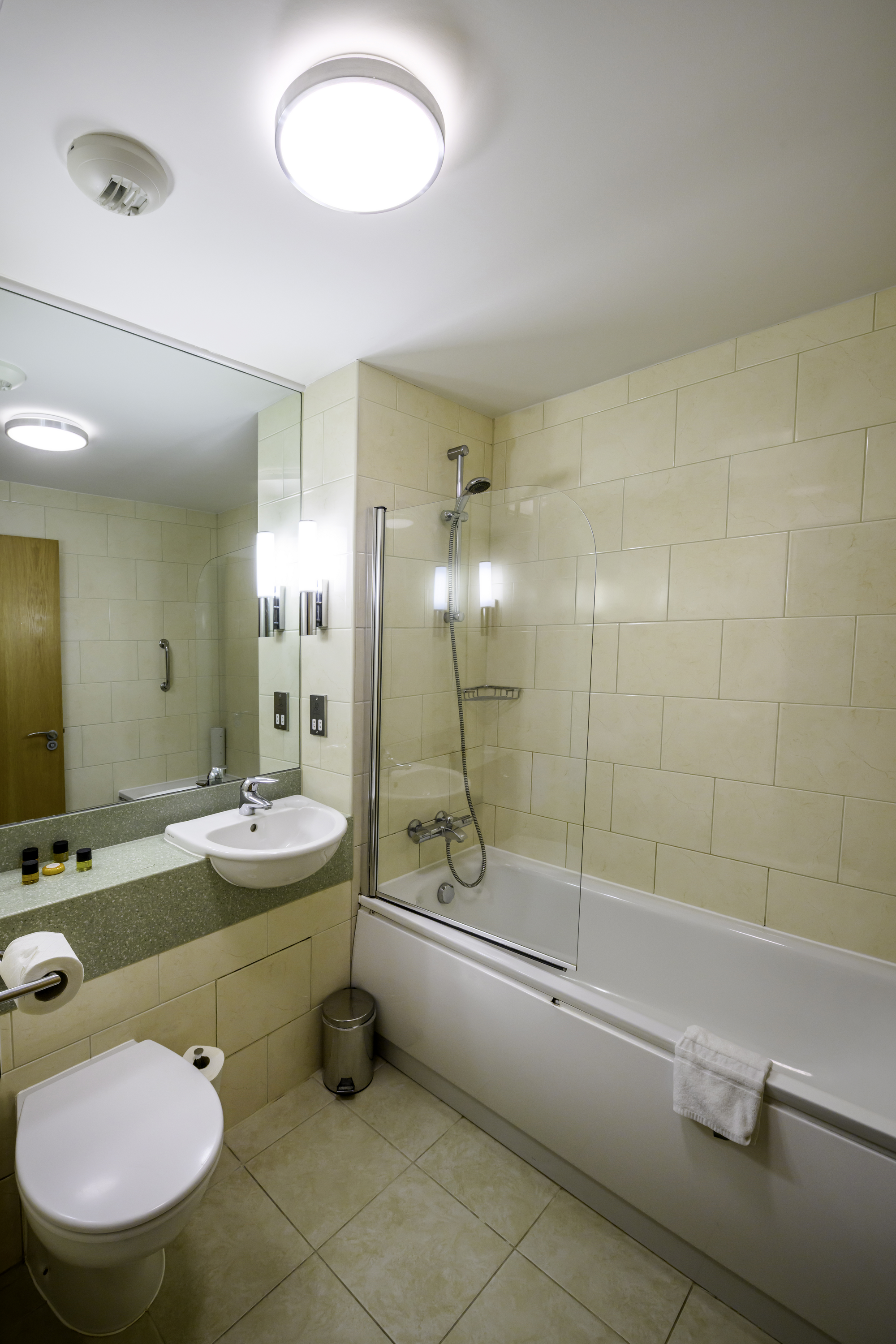 A modern bathroom with a bathtub, shower, toilet, and sink. The walls are tiled in light beige, with a large mirror above the sink. The ceiling has two circular light fixtures and a smoke detector.