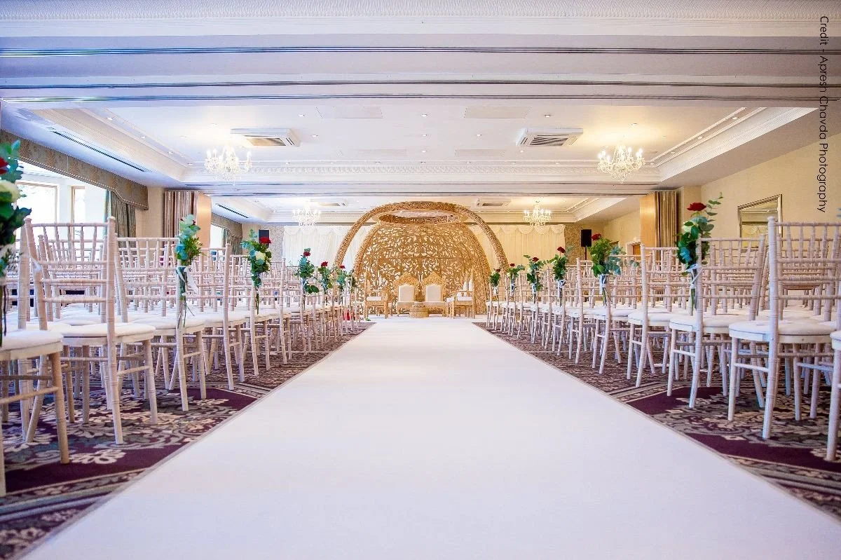 Elegant wedding ceremony setup with rows of white chairs decorated with red and white roses along a white aisle leading to a golden backdrop and two chairs.