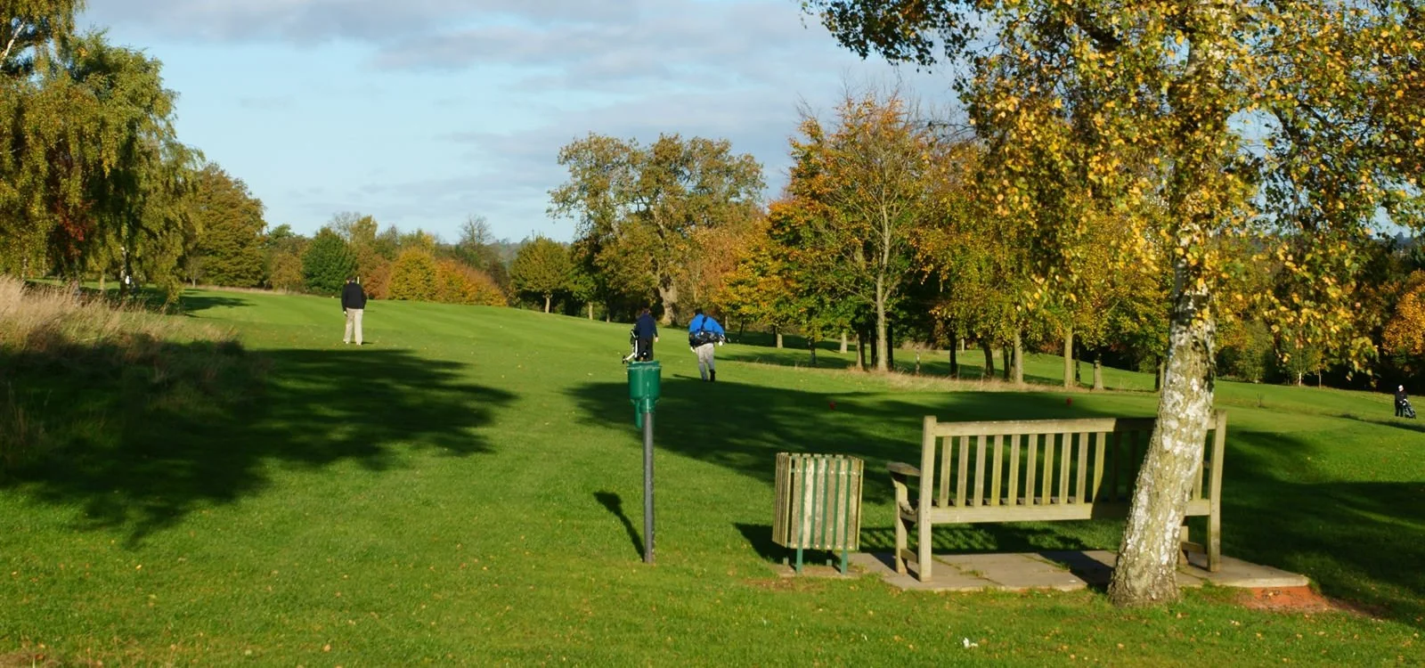A golf course with three golfers in the distance, surrounded by trees with fall foliage, a bench, and a trash bin in the foreground.