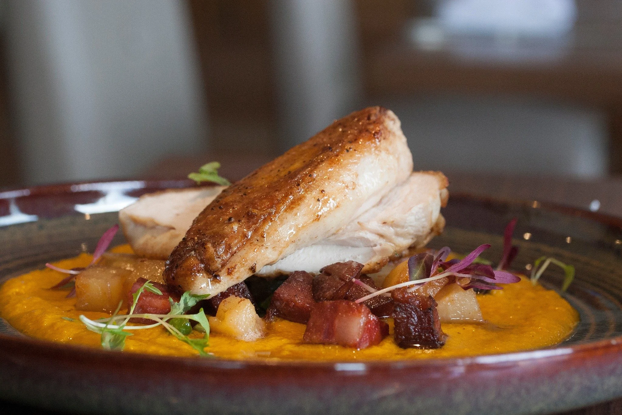 A plated dish featuring cooked chicken breast on top of a bed of diced vegetables and garnished with microgreens, served on a dark ceramic plate with a colorful glaze.