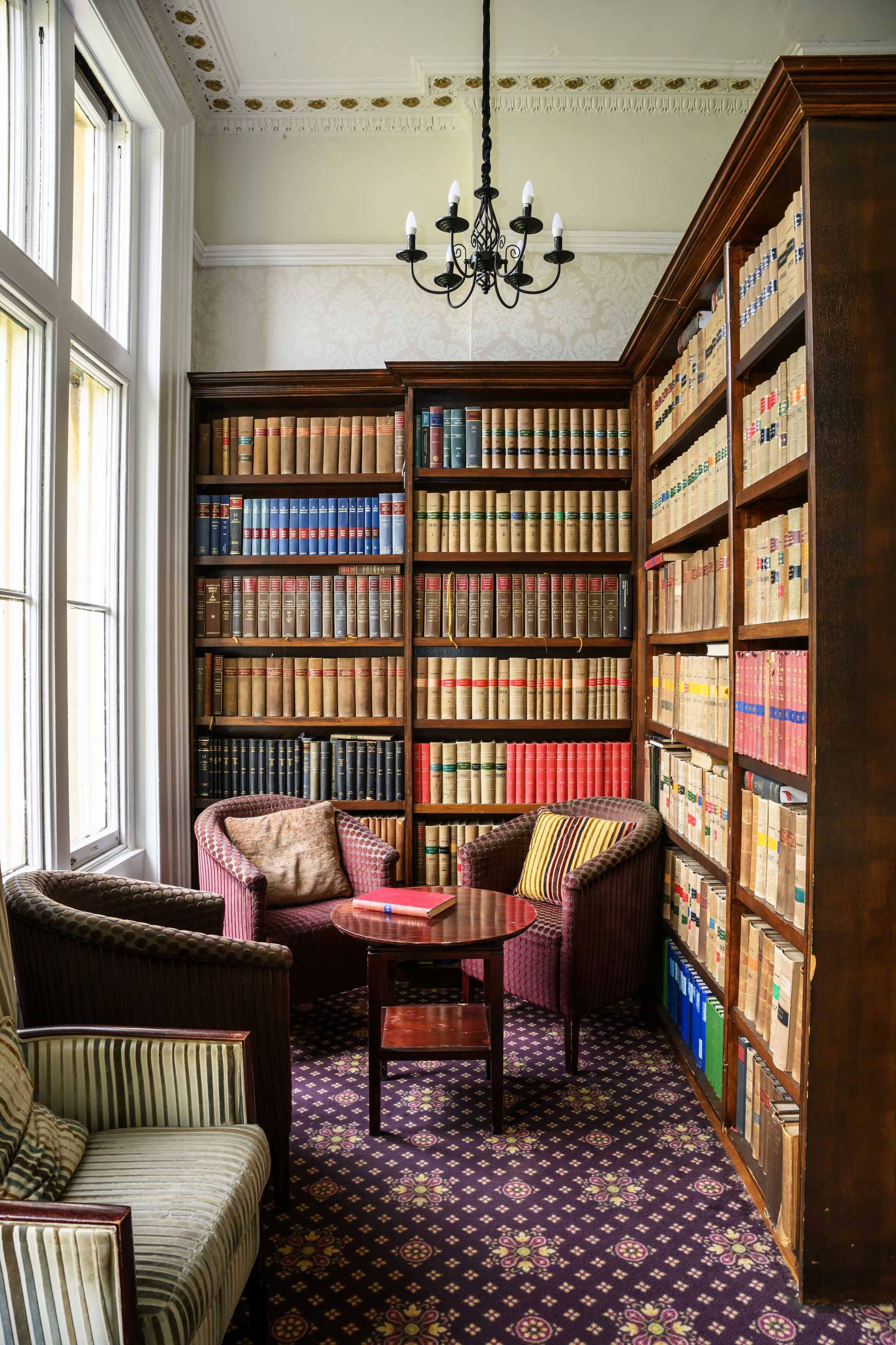 A cozy corner in a library with large windows, wooden bookshelves filled with books, a chandelier, a small round table, and upholstered chairs with patterned and striped pillows, beneath a decorative ceiling.