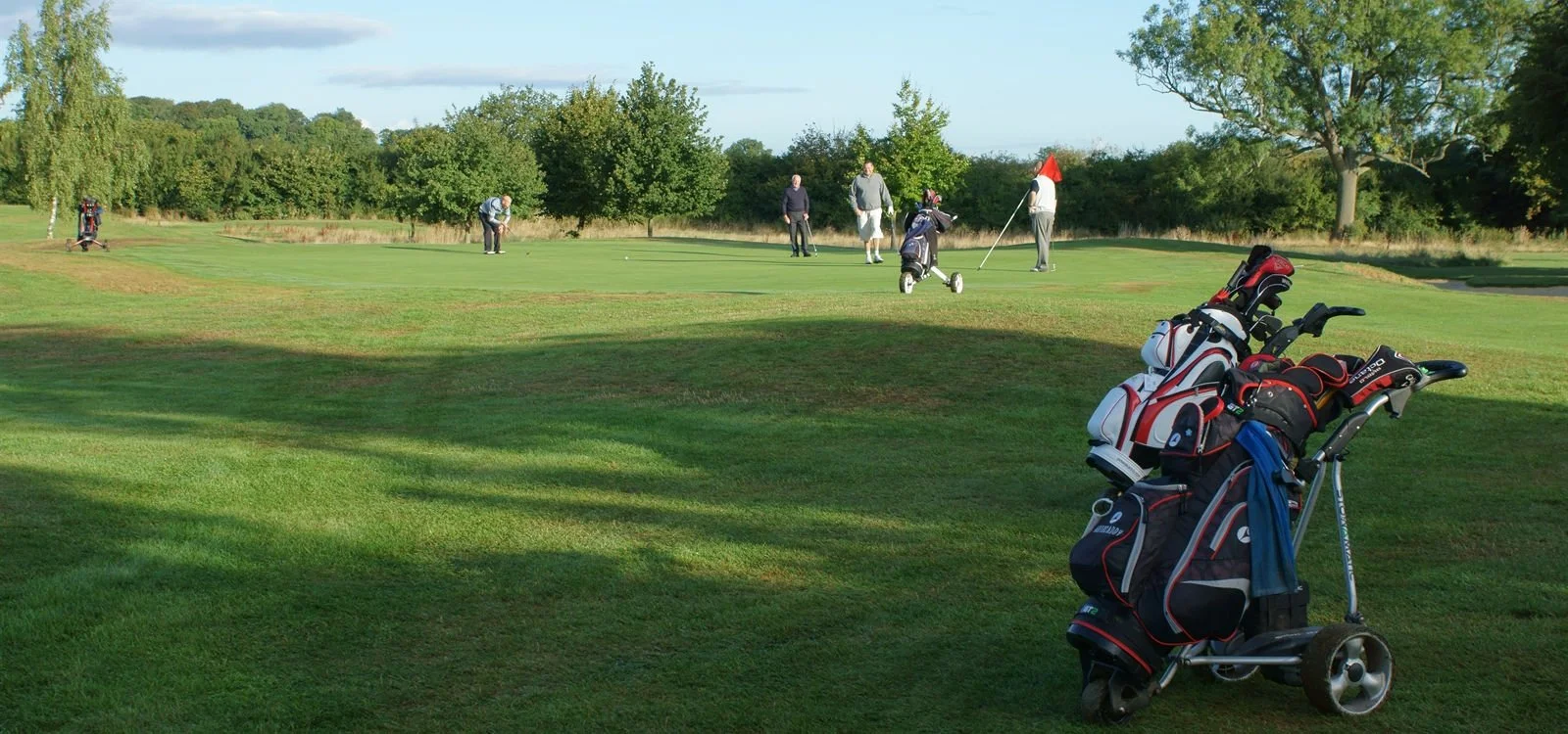 A golf course with five people playing golf near a green, with golf bags and clubs in the foreground, and trees in the background under a partly cloudy sky.