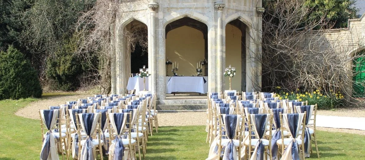 Outdoor wedding ceremony setup with rows of chairs facing an altar on steps, in front of a historic stone building with arches, surrounded by trees and bushes.
