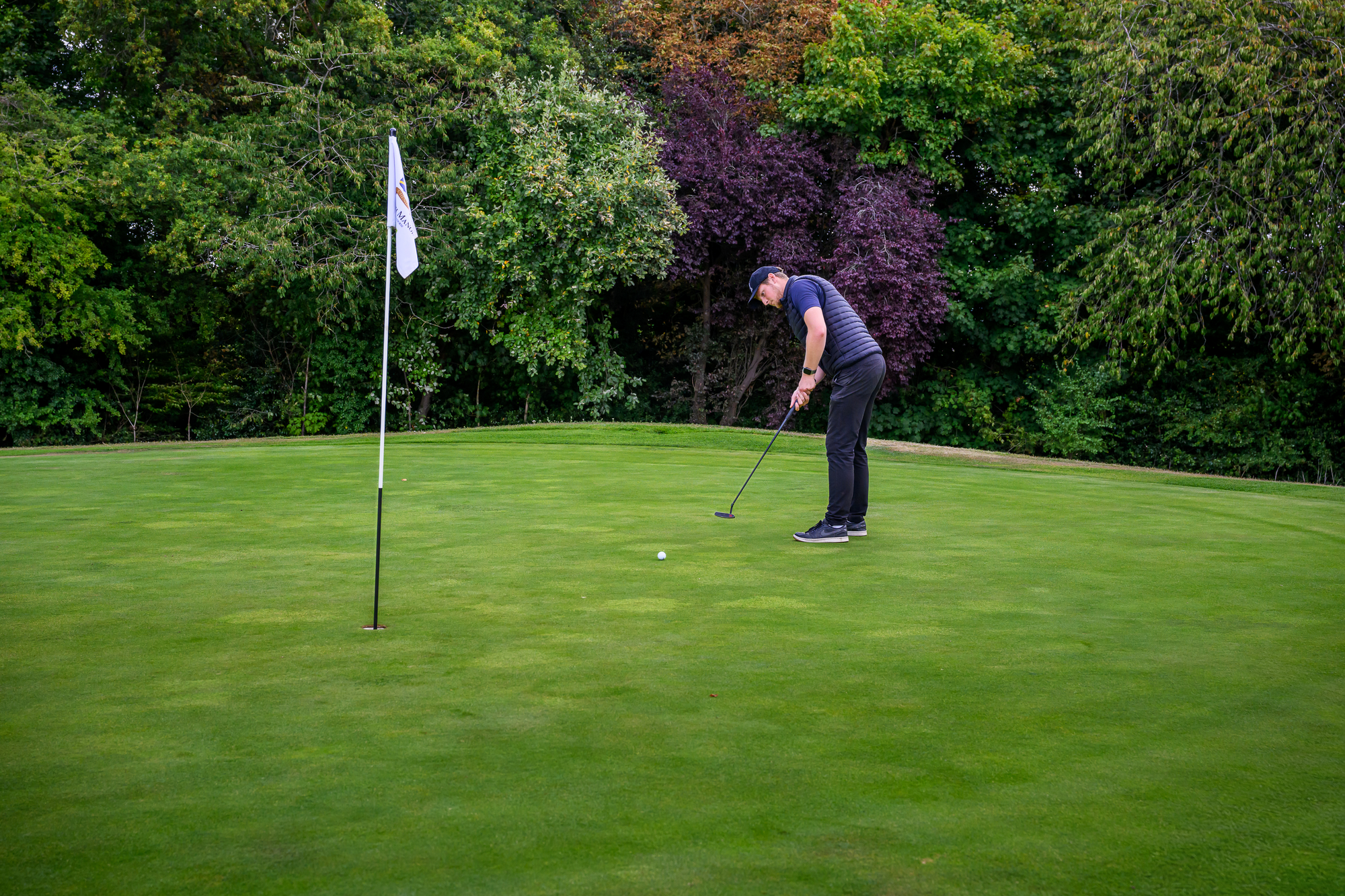 A man preparing to putt on a golf course green, with a flagstick nearby and trees in the background.