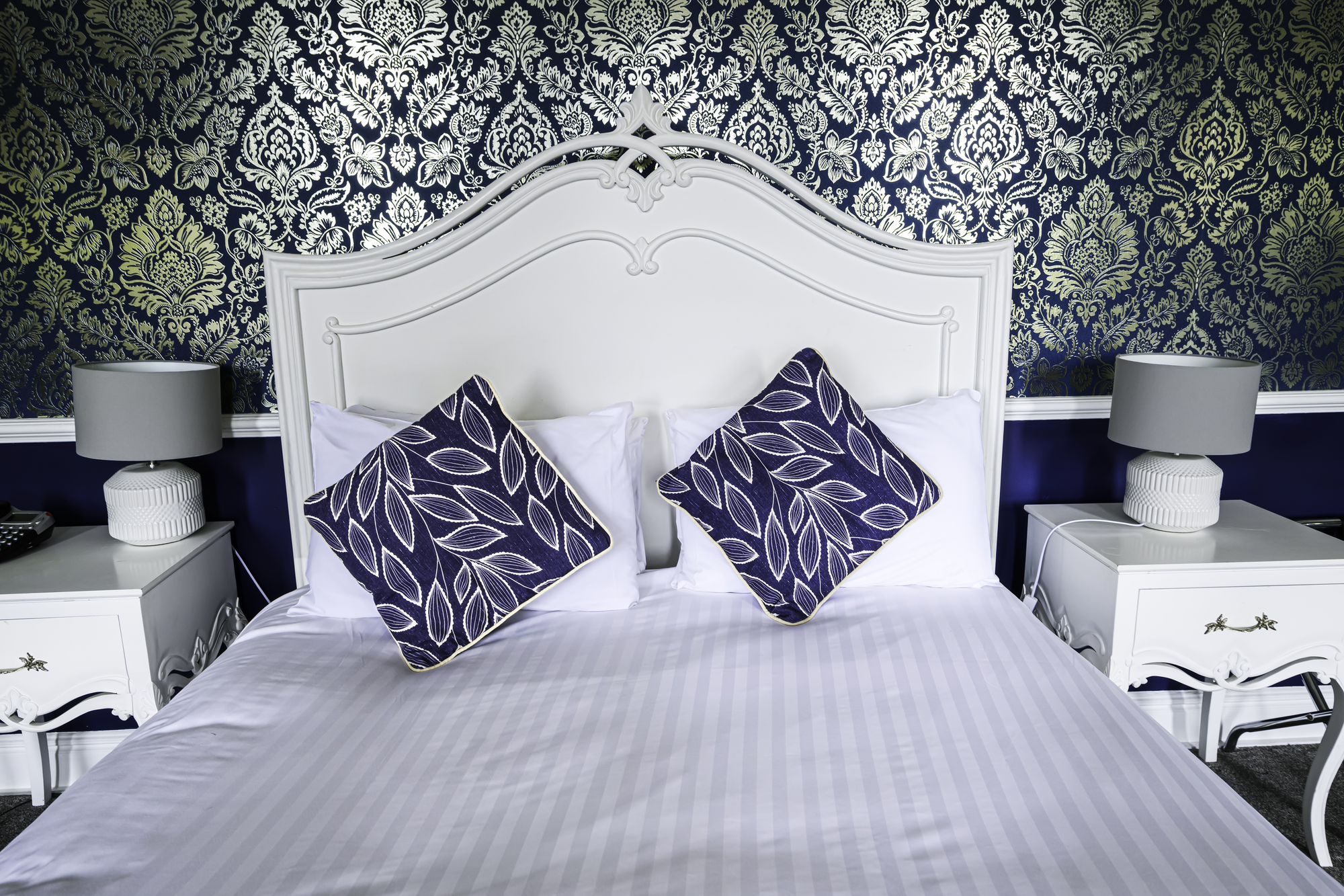 A white bed with two large pillows, each with navy and white leaf-patterned pillowcases, against a dark blue and gold damask-patterned wallpaper. There are two white nightstands, each with a gray lampshade, on either side of the bed.