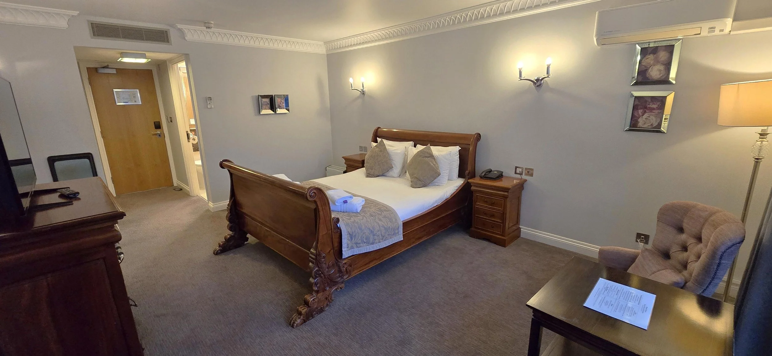 A hotel room with a wooden bed, beige bedding, and decorative pillows. There is a nightstand, a sitting chair, a desk with a remote control and television, and wall-mounted lights. An air conditioning unit is above the bed, and a doorway leads to a b