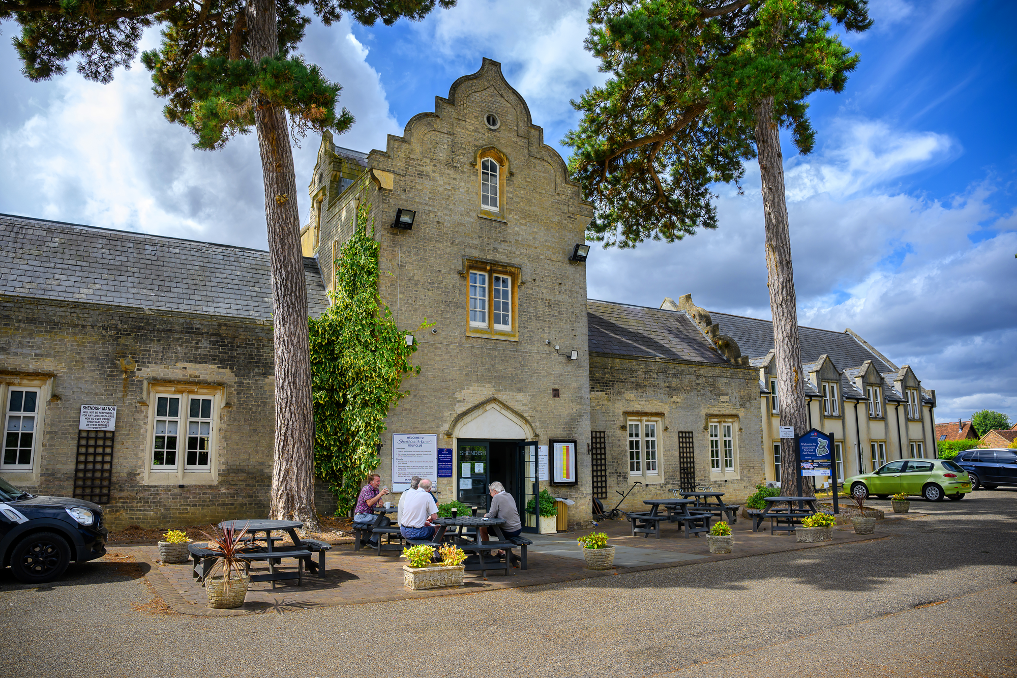 People sitting at outdoor tables in front of a historic stone building with trees and a cloudy sky.