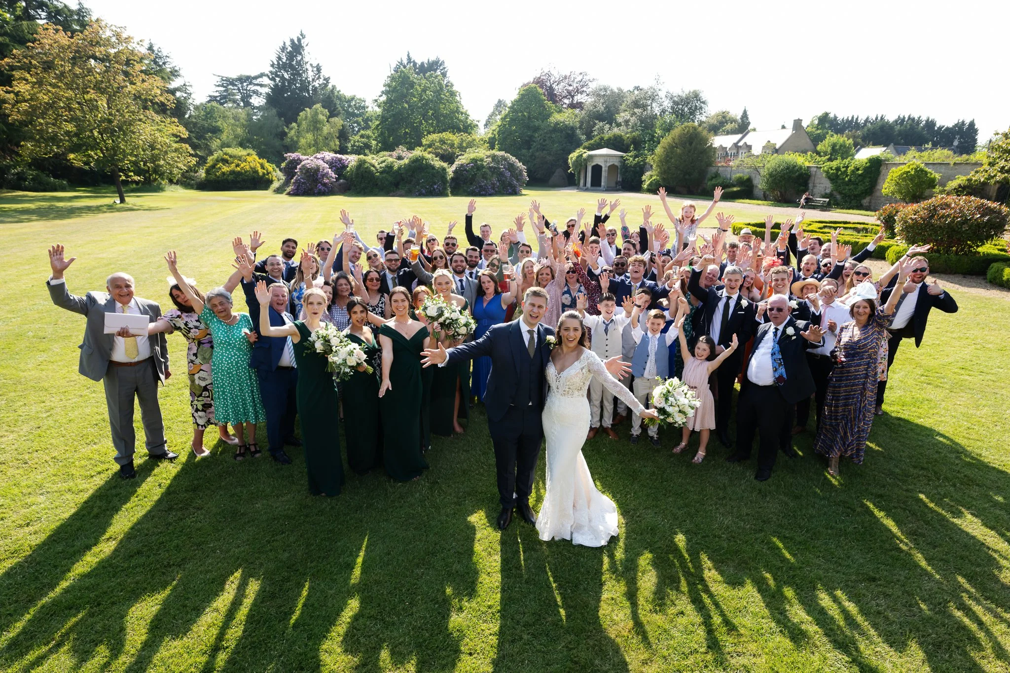 A large group of people, including a bride and groom, celebrating outdoors on a sunny day, with everyone raising their hands and smiling in a lush green park.