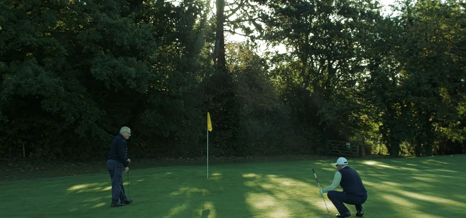 Two men are on a golf course, one standing and one crouching near a golf hole with a flag, preparing for putt, surrounded by trees.