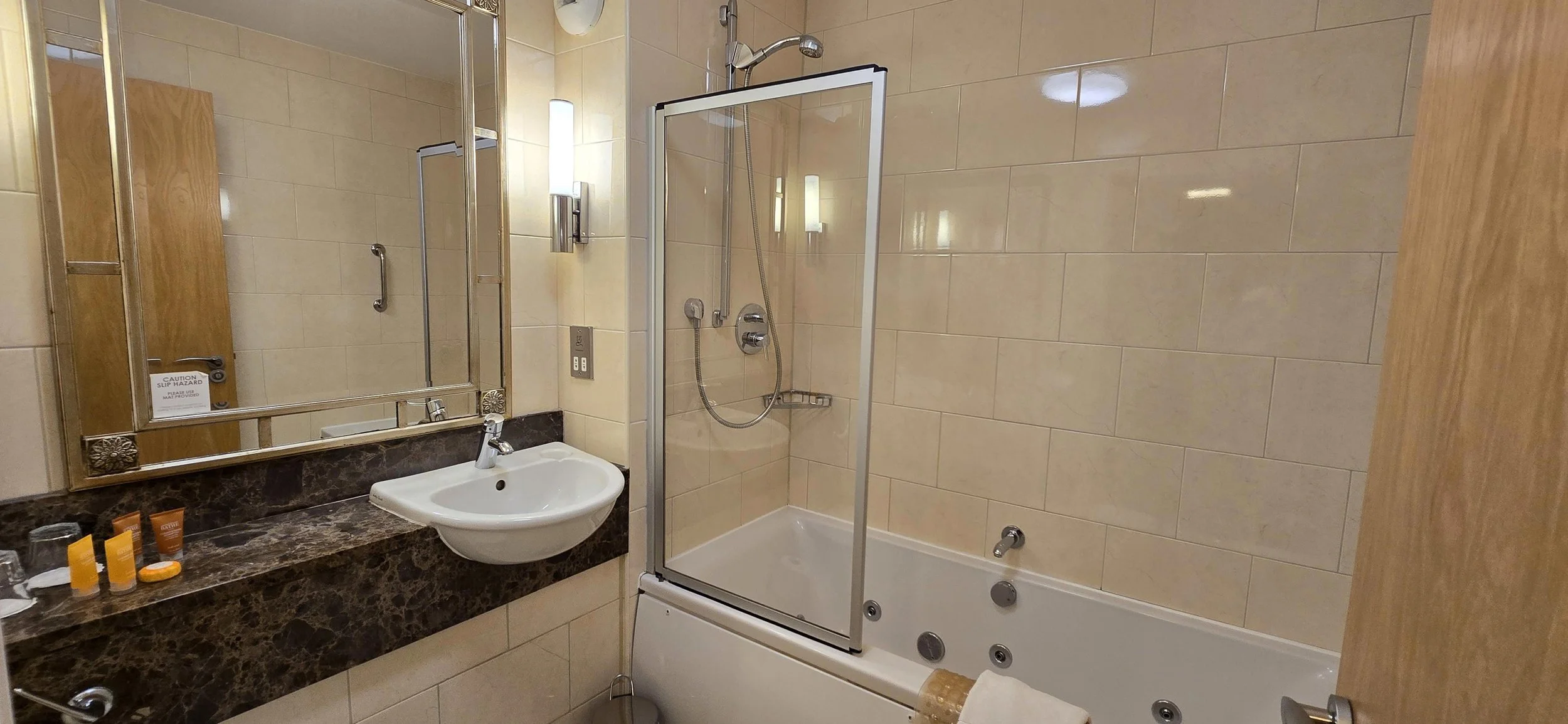 Bathroom with a shower, bathtub, and mirror. The shower has a glass door and handheld showerhead. The counter holds toiletries, and the room has beige tiles on the walls.