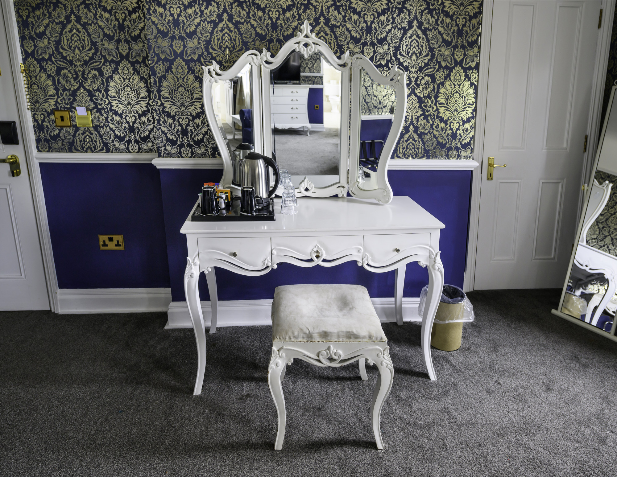 A white ornate makeup or vanity table with mirror, accompanied by a matching stool, in a room with dark blue and gold damask patterned wallpaper and dark carpet flooring. The table has a kettle, coffee mugs, and glasses on it. A door and a standing m